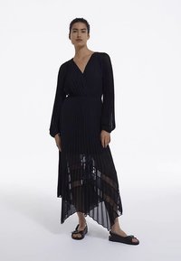 Černé plisované maxi šaty s výstřihem do V, dlouhými rukávy a průsvitnými spodními panely. Nošeny s černými sandály na nízkém podpatku, zdůrazňujícími strukturovanou látku.