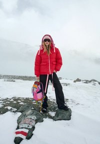 Czerwona puchowa kurtka z różowymi akcentami, czarne spodnie o szerokich nogawkach oraz wielokolorowy plecak. Zaśnieżony teren z kamienistym tłem i mgłą.