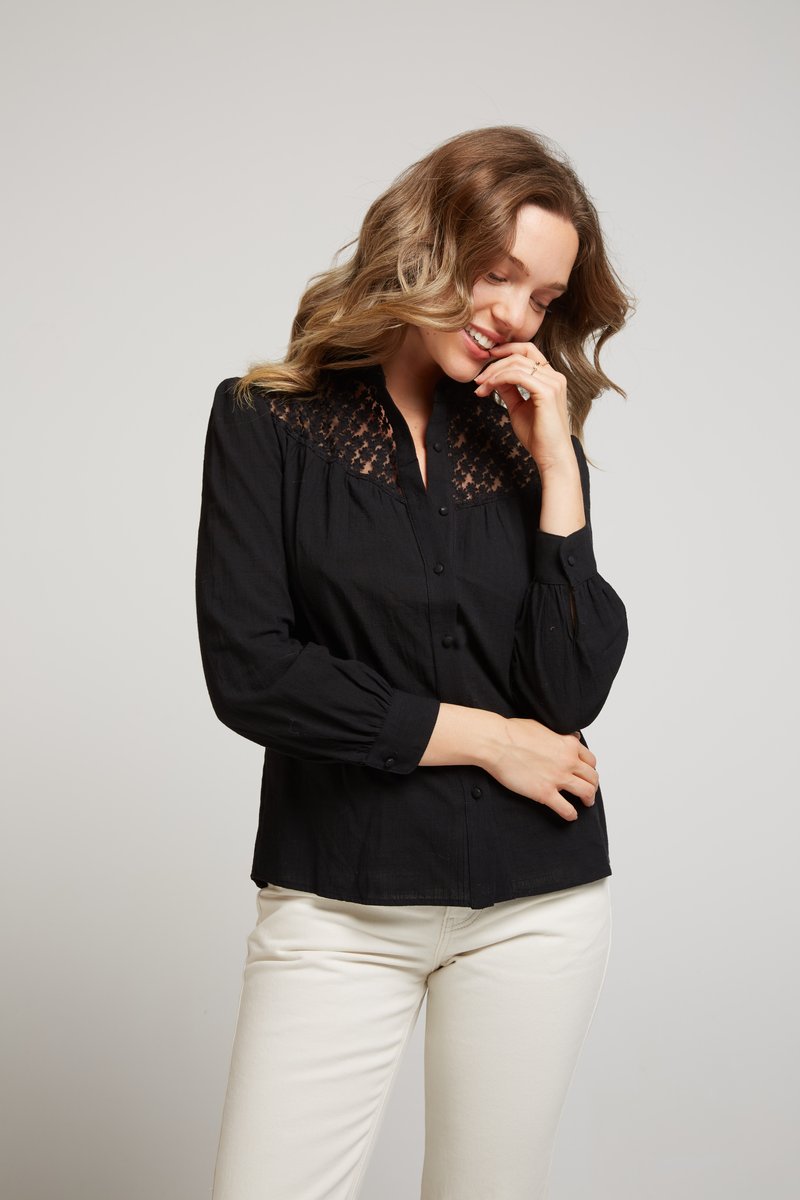 Blouse noire avec encolure en dentelle, longues manches bouffantes, fermeture à boutons et tissu doux. Assortie avec un pantalon blanc cassé pour le contraste.