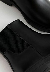 Des bottines noires en cuir présentent des panneaux latéraux élastiques, une finition lisse et une semelle plate, alliant design élégant et détails fonctionnels.