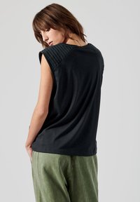 Haut sans manches noir avec des accents texturés aux épaules et une coupe ample, associé à un pantalon vert avec des clous décoratifs sur le côté.