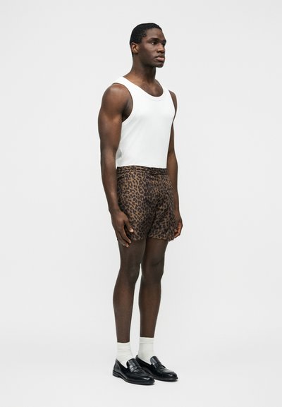 Homme debout portant un débardeur blanc, un short marron à motif léopard, des chaussettes blanches et des mocassins noirs, sur un fond uni.