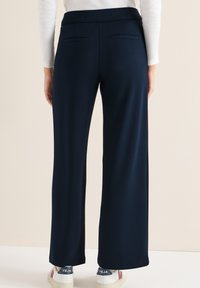 Personne portant un pantalon large bleu marine et un haut blanc à manches longues, vue de dos, debout contre un fond clair.