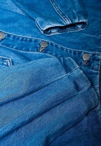 Zbliżenie na złożoną niebieską tkaninę dżinsową z widocznymi metalowymi guzikami i detalami szwów na kurtce lub spodniach dżinsowych.