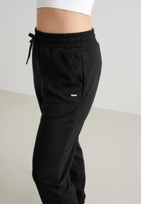 Schwarze sportliche Jogginghosen mit elastischem Bund und verstellbarem Kordelzug. Glatte Textur, seitliche Taschen und kleines silbernes Logodetail am Bein.