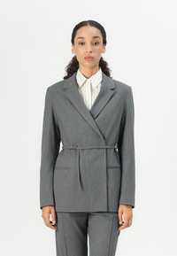 Grå dressjakke med pinstriper og skreddersydd passform, med snipede slag og detalj med belte på siden. Kombinert med en lysstripet skjorte med knapper.