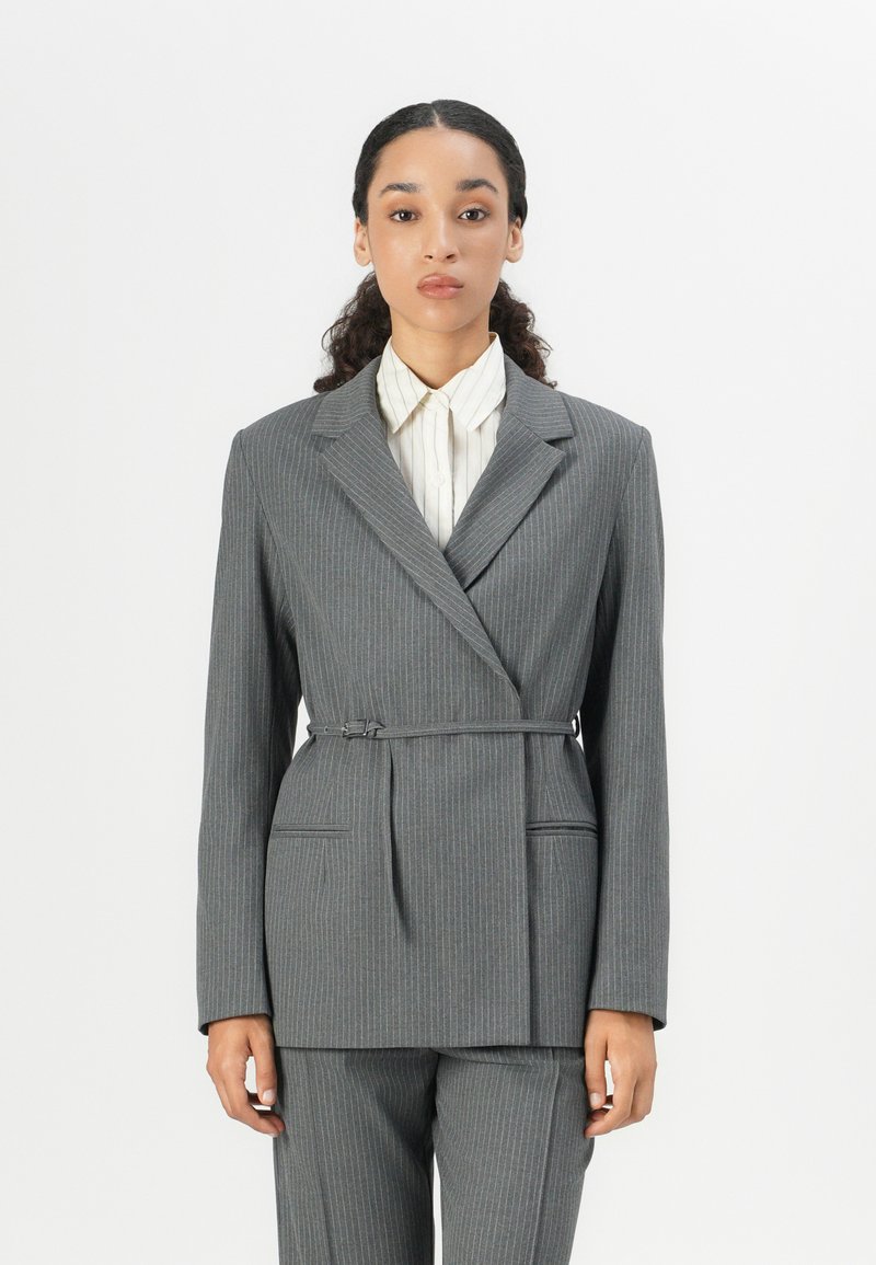Grå dressjakke med pinstriper og skreddersydd passform, med snipede slag og detalj med belte på siden. Kombinert med en lysstripet skjorte med knapper.