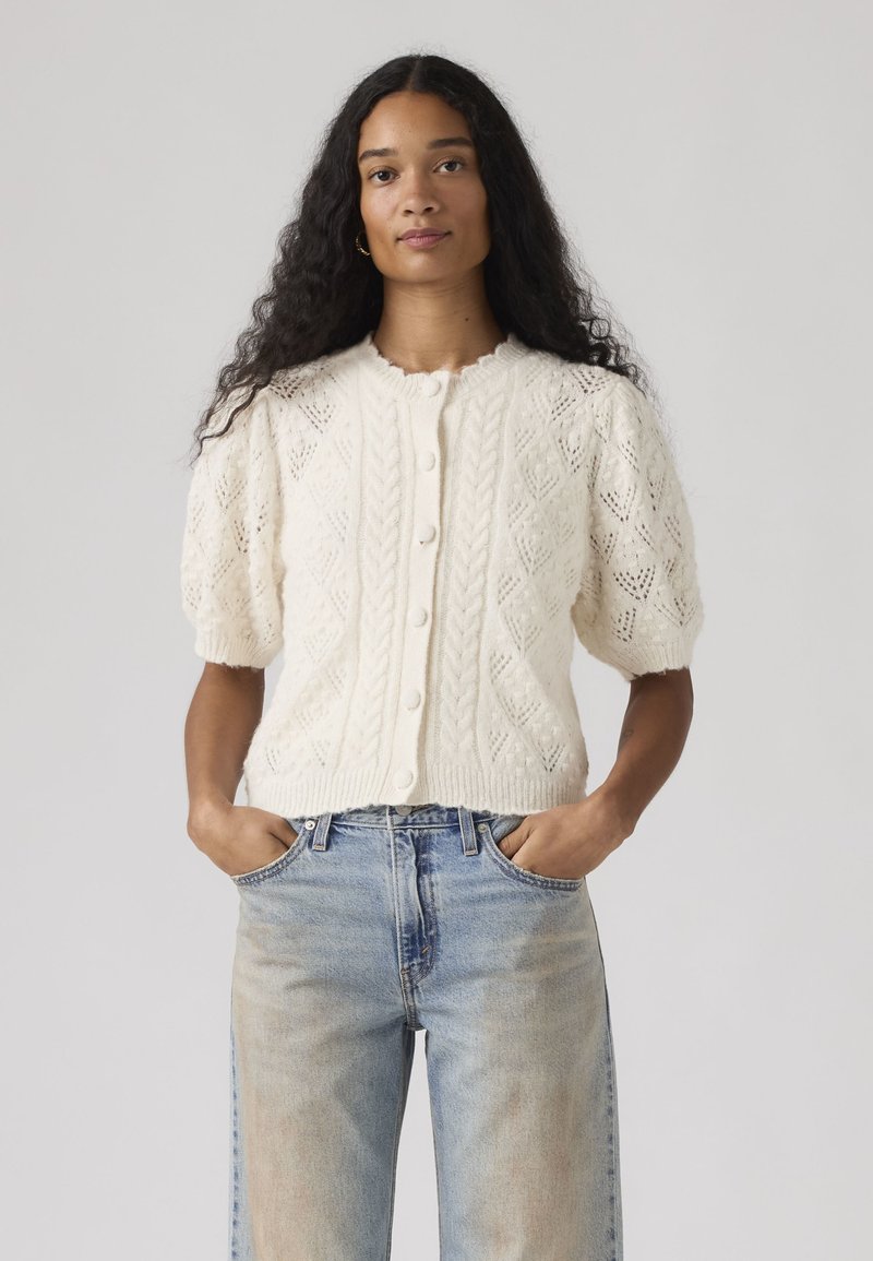 Woman with long curly hair wearing a white knitted short-sleeve buttoned cardigan and light blue jeans, hands in pockets.