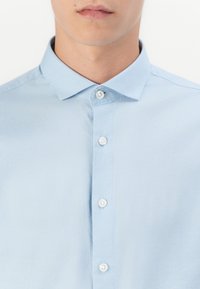 Man in een lichtblauw geknoopt overhemd met een klassieke kraag, weergegeven van de nek tot het midden van de torso tegen een witte achtergrond.