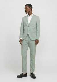 Traje verde claro con una chaqueta ajustada y pantalones ceñidos. Camisa blanca por debajo, complementada con mocasines negros y calcetines visibles hasta los tobillos.