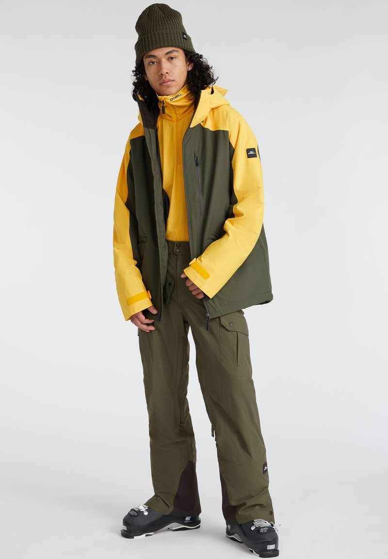 Skijacke in grün und gelb, mit durchgehendem Reißverschluss und Taschen, kombiniert mit olivfarbenen Hosen und schwarzen Skistiefeln, alles vor einer schneebedeckten Kulisse.