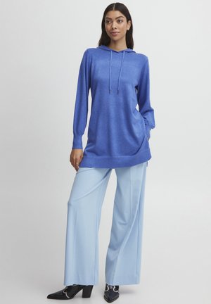 Blaues Kapuzenoberteil mit langen Ärmeln und geradem Saum, kombiniert mit hellblauen weiten Hosen und schwarzen Absatzstiefeln. Glatte Textur.