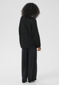 Een persoon met krullend haar staat met de rug naar ons toe in een zwart gespikkeld brei-jersey en losse zwarte wijde broek.
