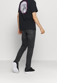 Persona alejándose con una camiseta negra que tiene un logo blanco bordeado en rosa, jeans gris oscuro y zapatillas negras con suelas blancas.