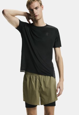 Bărbat blond purtând o cămașă neagră cu mâneci scurte și pantaloni scurți în straturi de culoare verde măsliniu, pozând cu mâna stângă în spatele gâtului, pe un fundal simplu.