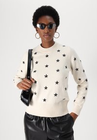 Pull crème avec des motifs d'étoiles noires, col rond, poignets et ourlet côtelés. Assortie à un pantalon en cuir noir et un sac à main noir.