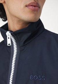 Homme portant une veste bleu marine avec une fermeture éclair blanche et "BOSS" brodé sur la poitrine, visible du cou jusqu'au milieu de la poitrine.