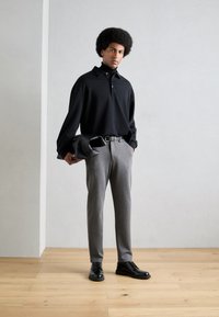 Črna majica polo z dolgimi rokavi in ovratnikom, sive krojene hlače in črni čevlji z vezalkami. Drži črno torbico.