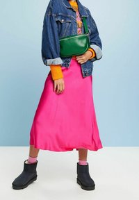 Veste en denim, pull orange vif, jupe midi en satin rose, sac bandoulière vert et bottes noires à semelle texturée.