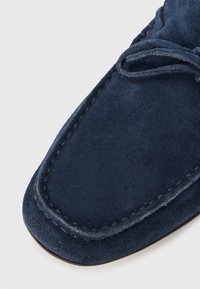 Mocassin en daim bleu marine avec une texture douce, un bout arrondi, des coutures décoratives et un détail de lacet contrastant sur la tige.
