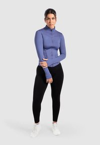 Lila Sportjacke mit Reißverschluss, tailliertem Schnitt, langen Ärmeln und Daumenlöchern, kombiniert mit schwarzen Leggings und weißen Turnschuhen.