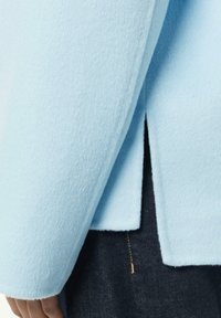 Giacchino blu chiaro in miscela di lana con una texture morbida, caratterizzato da un design con schiena spaccata. Indossato sopra jeans scuri con un dettaglio di cucitura dorata visibile.