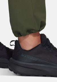 Zwarte Gore-Tex sneaker met een gestructureerd bovenwerk en dikke rubberen zool, gecombineerd met groene, opgerolde broekspijpen. Heeft een logo aan de zijkant.