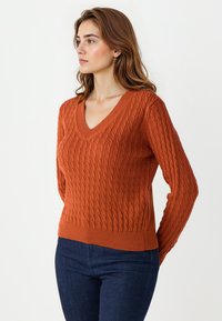 Pull en maille torsadée à col en V de couleur orange, avec des finitions côtelées et des manches longues, porté avec un jean bleu foncé. La texture est en relief et à motifs.
