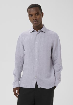 Mand iført en lysegrå langærmet skjorte med knapper og sorte bukser, der står foran en ensfarvet lys baggrund.