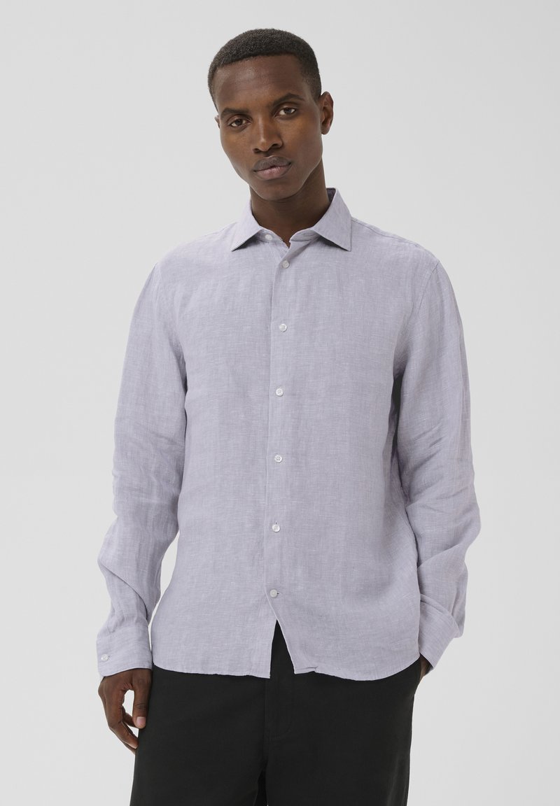 Homme portant une chemise à manches longues gris clair boutonnée et un pantalon noir, debout devant un fond clair uni.