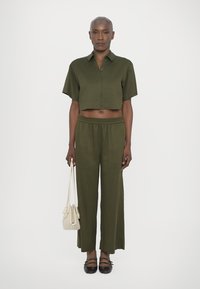 Chemise courte olive avec des manches courtes, associée à un pantalon ample olive. Les accessoires comprennent un sac à main léger et des chaussures noires.