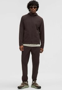 Braune Zip-Jacke und passende Hose aus leichtem Material. Verfügt über eine Kapuze, ein schlankes Design und elastische Bündchen. Kombiniert mit beigen und olivfarbenen Sneakern.