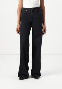 Jean droit en denim noir avec poches avant, porté avec des chaussures pointues noires ornées de détails en œillets argentés.