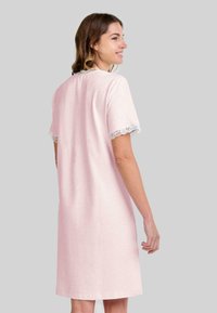 Mujer sonriente, vistiendo un vestido de corte a la rodilla de color rosa claro con bordados de encaje en las mangas y el cuello, de pie frente a un fondo gris liso.