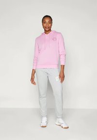 Ροζ φούτερ με λογότυπο μπροστά, σε συνδυασμό με γκρι φόρμες που έχουν λευκή λεπτομέρεια. Το μοντέλο στέκεται σε ανοιχτό φόντο, φορώντας λευκά αθλητικά παπούτσια.