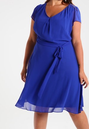 Sininen, polvipituinen mekko, jossa on lyhyet pussihihat, V-pääntie ja istuva vyötärönauha. Valmistettu kevyestä, laskeutuvasta kankaasta, jossa on sileä pinta.