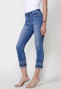 Jean en denim bleu avec coupe slim, comportant des motifs en diamant brodés et des ourlets à franges. Portés avec des sandales à talons blocs transparentes.