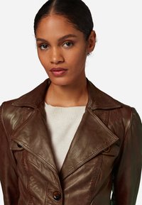 Brown leather jacket with a double-breasted design, pointed collar, and button accents. Worn over a light sweater, showing smooth texture.
