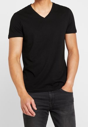 Mężczyzna w dopasowanej czarnej koszulce z krótkim rękawem i dekoltem w serek, z jedną ręką w kieszeni, ubrany w czarne dżinsy na jednolitym tle.