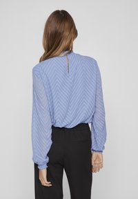 Blouse bleu clair à motif chevron avec des manches longues et froncées ; présente un décolleté en forme de trou de serrure et est associée à un pantalon noir taille haute.