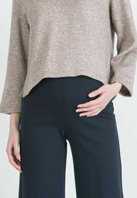 Beige, teksturoitu neulepaita rentoistuvalla mitalla ja 3/4 hihalla, yhdistettynä mustiin, leveälahkeisiin housuihin, joiden pinta on sileä.