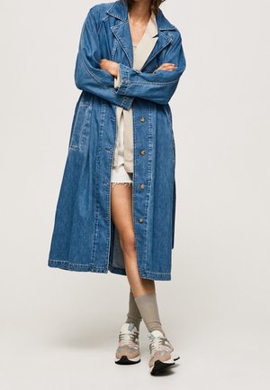 Persona vistiendo un largo abrigo vaquero azul sobre un conjunto beige con calcetines beige y zapatillas beige, blancas y azules, de pie con los brazos cruzados.