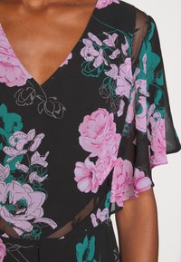 Femme portant une robe noire à col en V avec un motif floral rose et des manches transparentes flottantes laissant apparaître une partie du bras et de la poitrine.