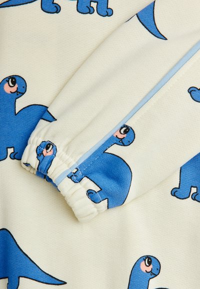 Motivi di dinosauri blu su un tessuto crema, con chiusura a zip e polsini arricciati. La texture è liscia e morbida.