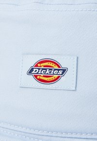 Lyseblå stoff med et rektangulært hvitt merke som har "Dickies"-logoen, som inkluderer røde og gule sirkulære detaljer.