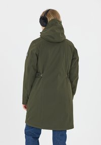 Olijfgroene parka met capuchon, voorzien van een gladde textuur, twee zijknopen en een iets losse pasvorm. Gemaakt met een spijkerbroek.