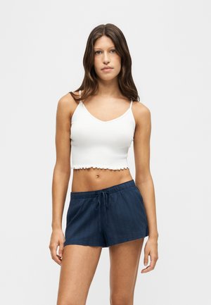 Jeune femme aux longs cheveux bruns portant un crop top blanc et un short bleu marine à cordon, debout devant un fond clair uni.