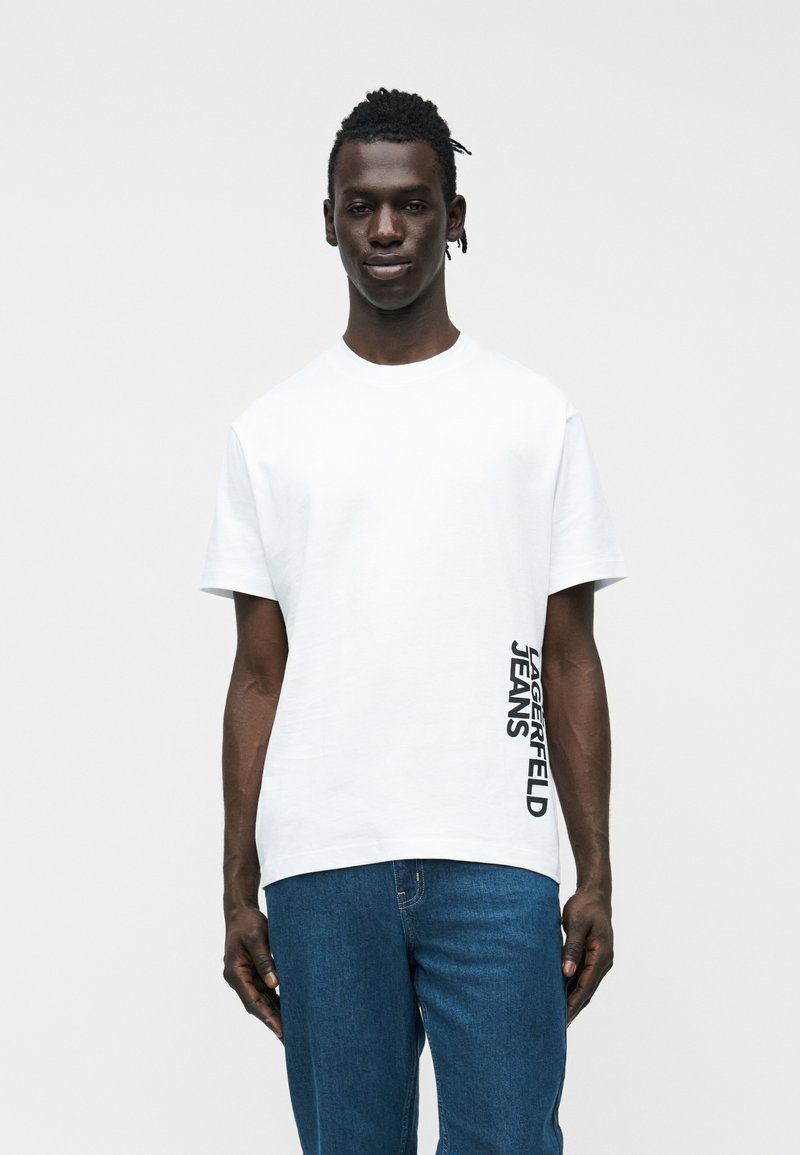 Άνδρας που φοράει λευκό T-shirt με κατακόρυφο κείμενο "JAEGERFELD JEANS" στο πλάι και μπλε τζιν παντελόνι, που στέκεται μπροστά σε απλό ανοιχτό φόντο.