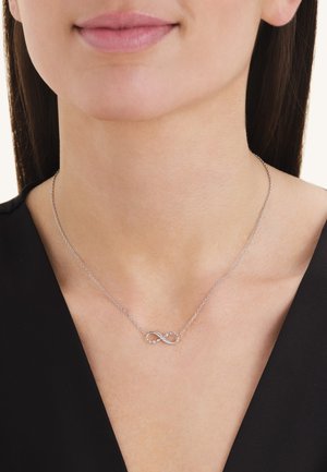 Delicate zilveren infinity-ketting met kleine ingelegde steentjes, gedragen door een vrouw met stijl donker haar en een zwart topje.