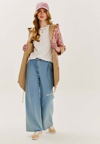 Dlhý kabát s kvetinovým vzorom a béžovým interiérom, biele tričko, svetlomodré široké džínsy a béžové tenisky, doplnené ružovou šiltovkou.